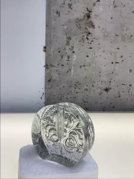 Deze solifleur naar een ontwerp van Heiner Deusterhaus uit de zeventiger jaren, is gemaakt van transparant glas en heeft aan een zijde een bloemmotief glad gesneden op een ruwe achtergrond.