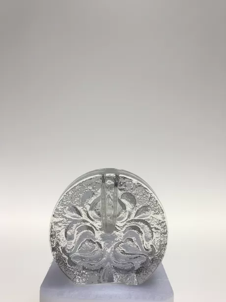 Deze solifleur naar een ontwerp van Heiner Deusterhaus uit de zeventiger jaren, is gemaakt van transparant glas en heeft aan een zijde een bloemmotief glad gesneden op een ruwe achtergrond.