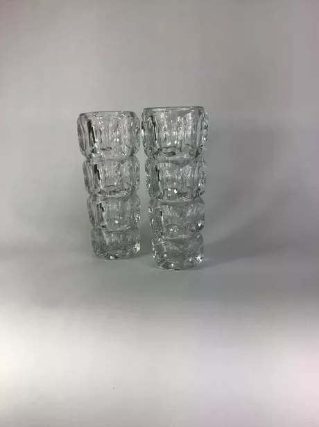 Twee Glazen Lensvazen František Pečený Deze uniek set naar een ontwerp van František Pečený (1920–1977) uit de jaren '60 vier fijn gesneden etages. Pečený was een toonaangevende Tsjechische ontwerper van geperst glas. Vanaf 1947 werkte hij als huisontwerper bij Heřmanova huť, waar hij decennia lang servies- en sierglas ontwierp: heldere, optisch spelende vormen met reliëf ("lens-/'thousand eyes'"-motieven) en in de jaren zeventig o.a. de bekende “candle wax”-vazen (model o.a. 20245, 1974), waarvan de onderhavige set een voorloper is.