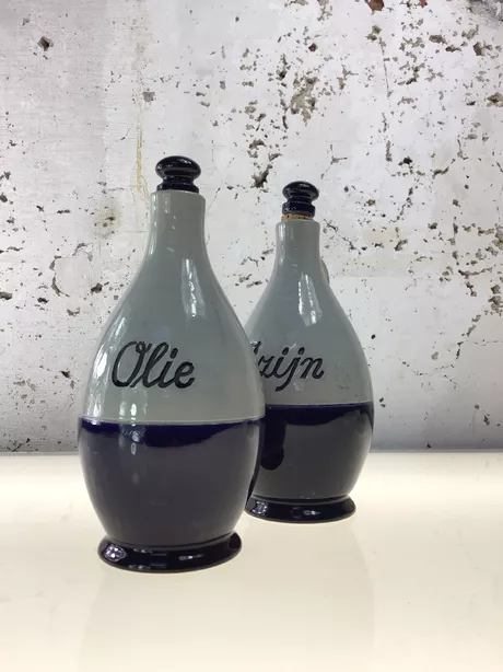 Een Olie-en-Azijn-kan van Reinhold Merkelbach, Höhr-Grenzhausen is typisch Westerwald steengoed: grijs zoutglazuur met kobaltblauwe penselering of reliëfdecor; vaak een slanke, cilindrische kruikvorm (Ø ca. 10 cm, H ca. 25 cm) met oor en email/kurk schroefdop. Deze set is gemerkt met “Reinhold Merkelbach Grenzhausen”.