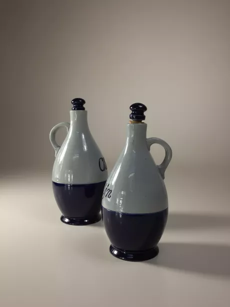 Een Olie-en-Azijn-kan van Reinhold Merkelbach, Höhr-Grenzhausen is typisch Westerwald steengoed: grijs zoutglazuur met kobaltblauwe penselering of reliëfdecor; vaak een slanke, cilindrische kruikvorm (Ø ca. 10 cm, H ca. 25 cm) met oor en email/kurk schroefdop. Deze set is gemerkt met “Reinhold Merkelbach Grenzhausen”.