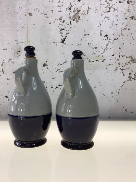 Een Olie-en-Azijn-kan van Reinhold Merkelbach, Höhr-Grenzhausen is typisch Westerwald steengoed: grijs zoutglazuur met kobaltblauwe penselering of reliëfdecor; vaak een slanke, cilindrische kruikvorm (Ø ca. 10 cm, H ca. 25 cm) met oor en email/kurk schroefdop. Deze set is gemerkt met “Reinhold Merkelbach Grenzhausen”.