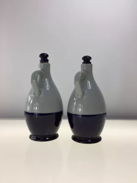 Een Olie-en-Azijn-kan van Reinhold Merkelbach, Höhr-Grenzhausen is typisch Westerwald steengoed: grijs zoutglazuur met kobaltblauwe penselering of reliëfdecor; vaak een slanke, cilindrische kruikvorm (Ø ca. 10 cm, H ca. 25 cm) met oor en email/kurk schroefdop. Deze set is gemerkt met “Reinhold Merkelbach Grenzhausen”.