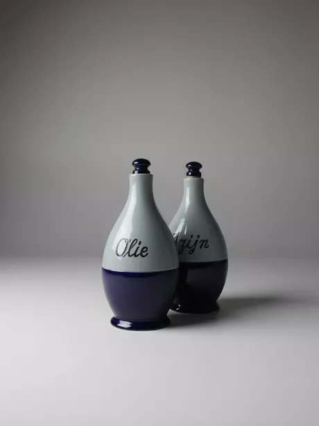 Een Olie-en-Azijn-kan van Reinhold Merkelbach, Höhr-Grenzhausen is typisch Westerwald steengoed: grijs zoutglazuur met kobaltblauwe penselering of reliëfdecor; vaak een slanke, cilindrische kruikvorm (Ø ca. 10 cm, H ca. 25 cm) met oor en email/kurk schroefdop. Deze set is gemerkt met “Reinhold Merkelbach Grenzhausen”.