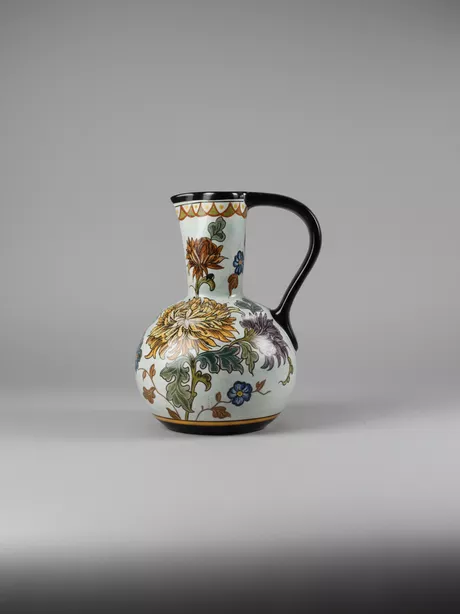 Plateel Schenkkan Vaas Chrysant thema Gouda Holland Deze fraaie schenkkan heeft rondom een bloemmotief (chrysanten met stengels en bladeren) op een lichtblauwe achtergrond. Onbeschadigd en puntgaaf.